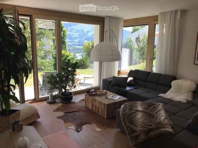 Living Room. Modern living room with large windows offering a scenic mountain view, featuring a black sectional sofa, a wooden coffee table, a floor lamp, and decorative plants.