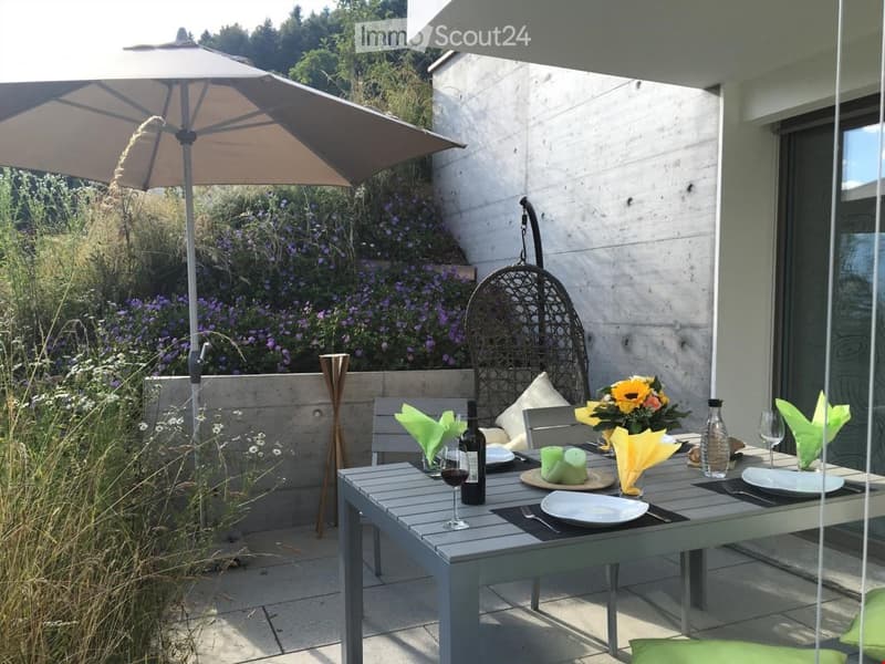 Garden / Outdoor Area. A modern outdoor dining area with a table set for two, surrounded by greenery and a hanging egg chair. The area features a large umbrella providing shade and is adjacent to a building with large windows.