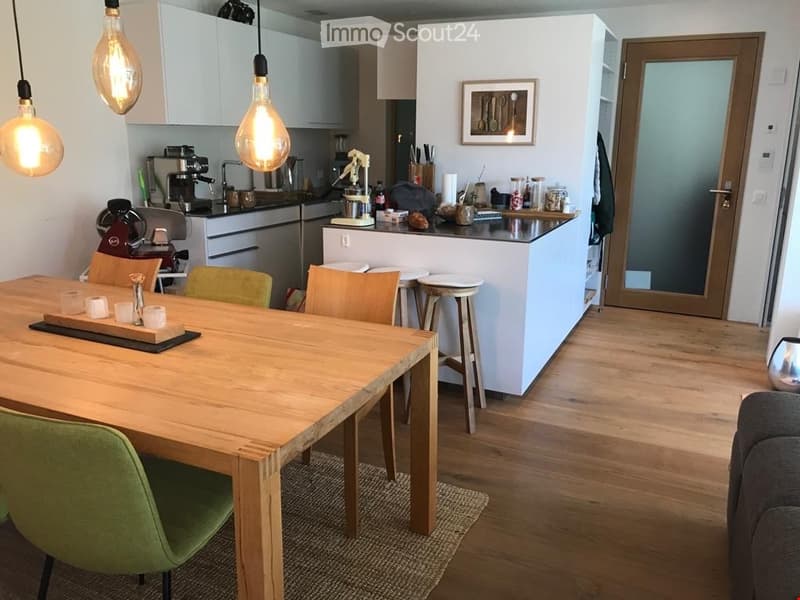 Furnished Room. A modern, open-plan kitchen and dining area with a wooden dining table, chairs, and a kitchen island. The kitchen features white cabinets, a coffee maker, and various appliances. The space is well-lit with hanging light bulbs and has a sle