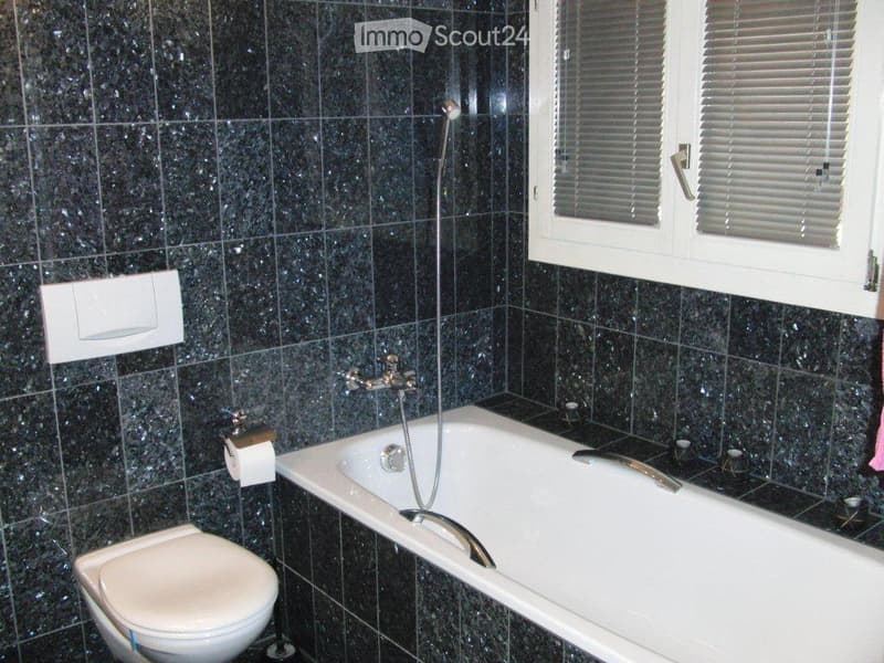 Bathroom with black tiled walls, white bathtub, showerhead, toilet, window with blinds