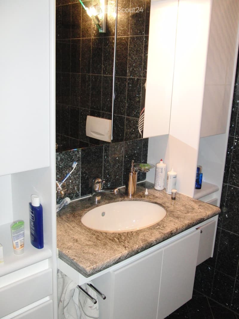 Bathroom with granite countertop, undermount sink, vanity cabinets, a mirror, shelves, and black tiled walls