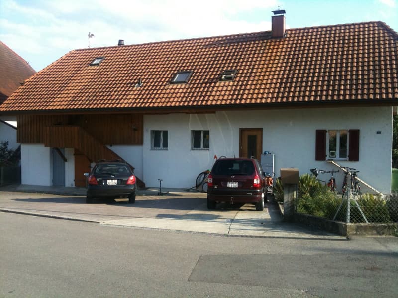 House. Two-story house with a red-tiled roof, white exterior walls, and a wooden balcony on the left side. The house has a garage with two parked cars, bicycles, and a small garden area with shrubs.