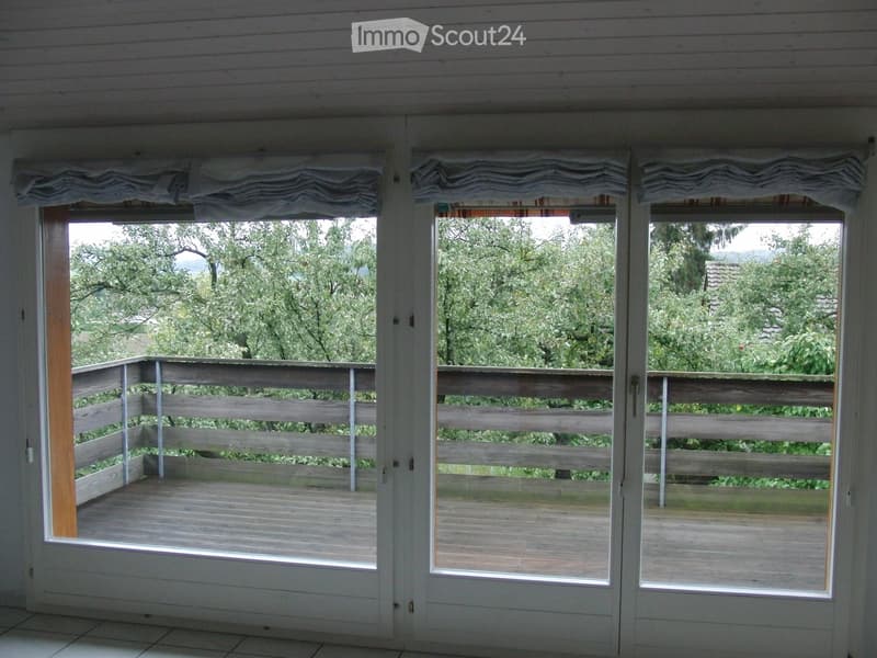 Balcony / Terrace. Large sliding glass doors open to a wooden balcony with a railing, overlooking a lush green garden and trees.