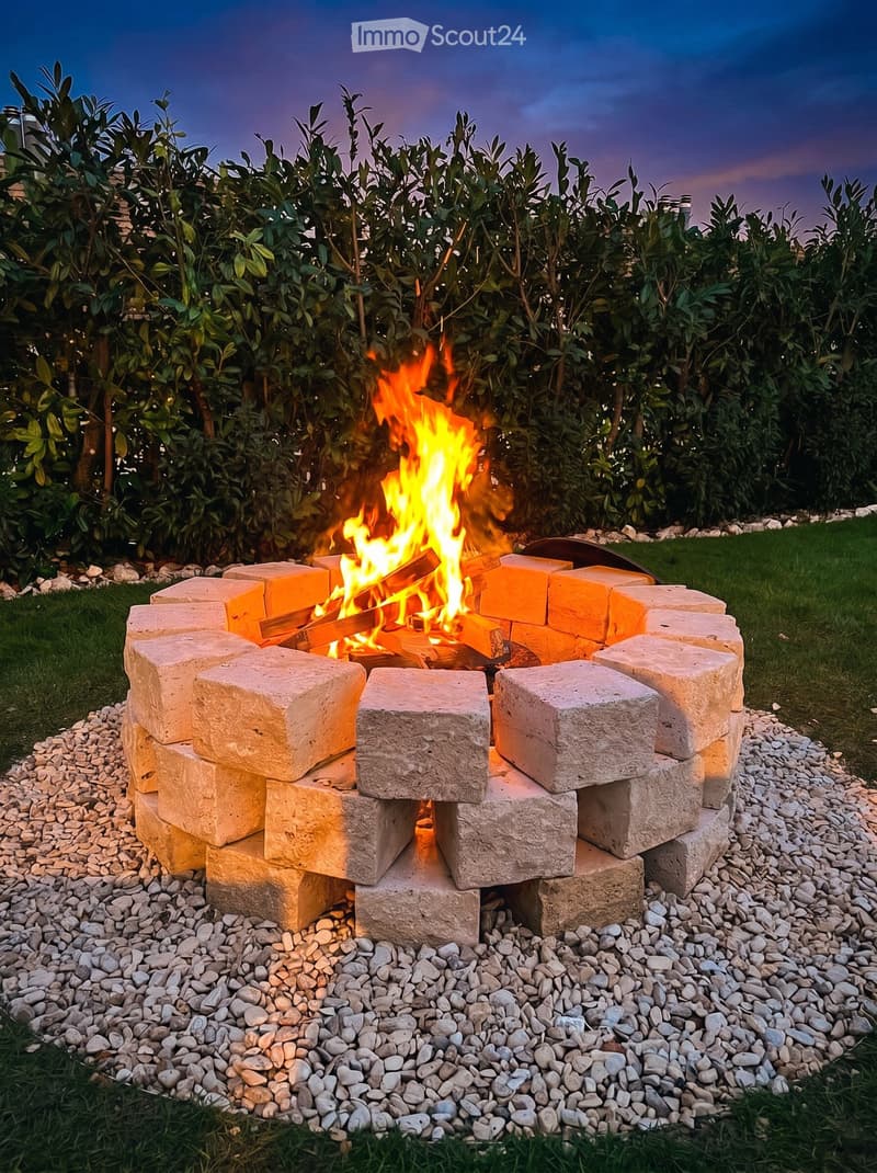 Garden / Outdoor Area. A stone fire pit with a roaring fire, surrounded by a bed of small rocks and lush green foliage.