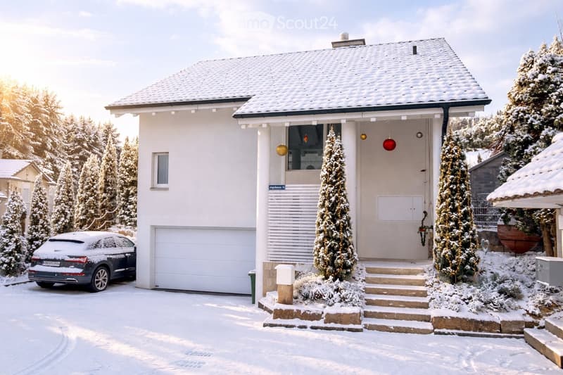 House. Two-story house with a snow-covered roof and garage, surrounded by snow-covered trees and a parked car.