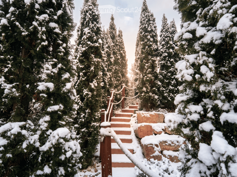Garden / Outdoor Area. A serene garden pathway lined with snow-covered evergreen trees, featuring wooden steps and a wooden railing leading up a gentle slope.
