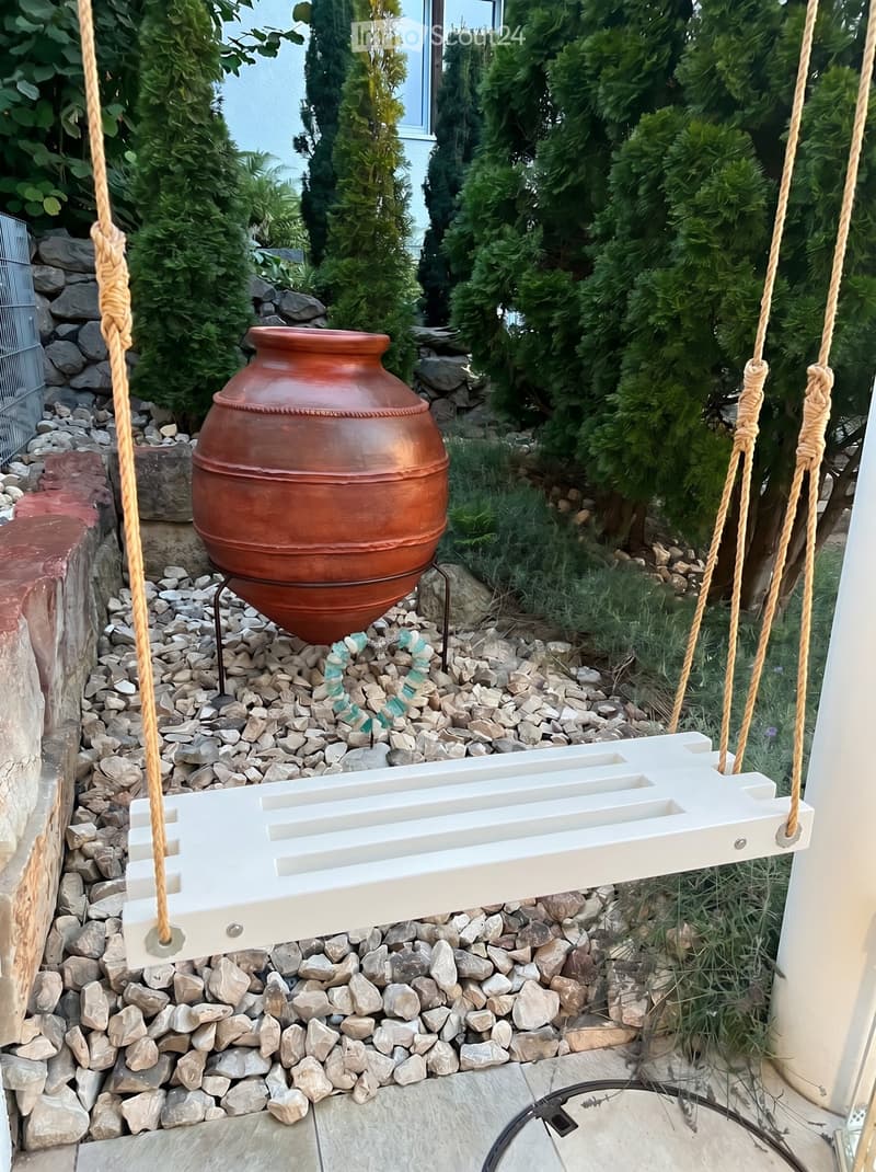 Garden / Outdoor Area. Garden area with a large terracotta vase on a stone base, surrounded by rocks and greenery, with a hanging white bench or swing in the foreground.