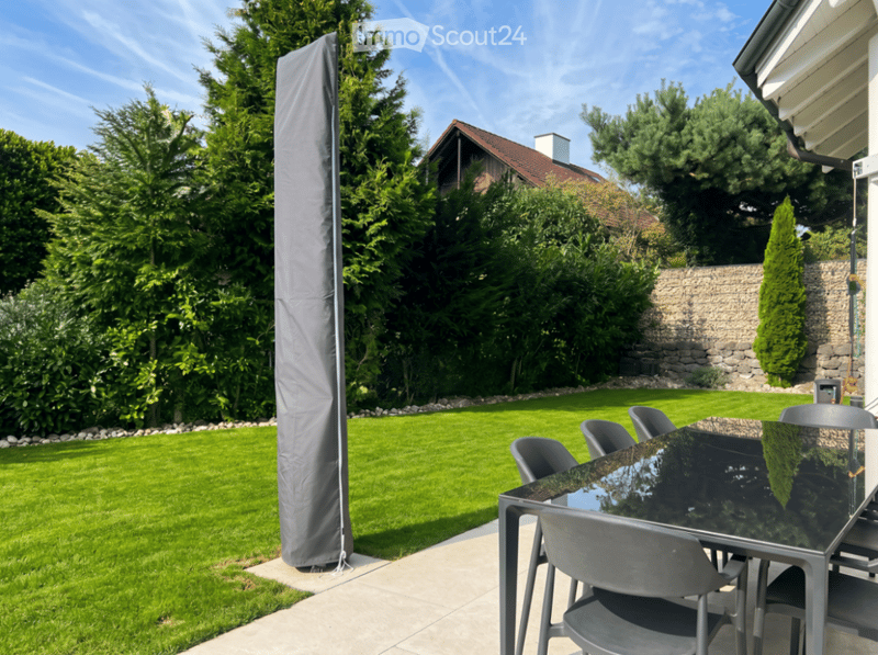 Garden / Outdoor Area. Well-maintained garden with a neatly trimmed lawn, tall trees, and a patio area featuring a glass-top dining table and chairs. A tall, grey outdoor umbrella is positioned near the lawn.
