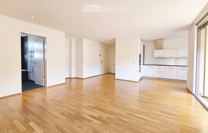 Unfurnished Room. Empty room with light wooden flooring, white walls, and a view into a kitchen area with white cabinets and appliances.