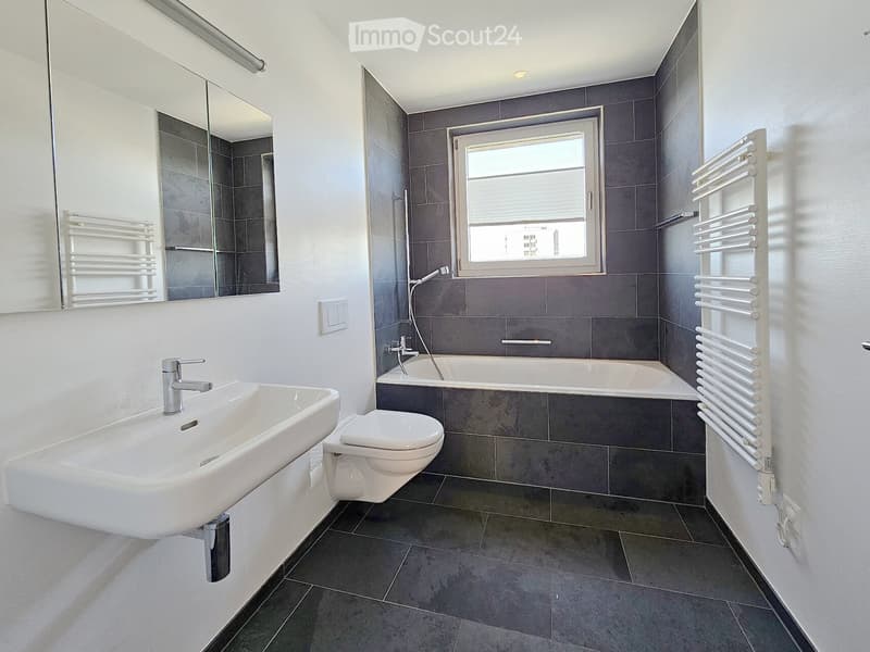 Bathroom. Modern bathroom with a white sink, toilet, bathtub, and large mirror. Grey tiled walls and floor, radiator on the right side, and a window above the bathtub.