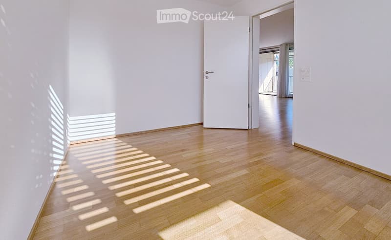 Unfurnished Room. Empty hallway with light wooden flooring and white walls, featuring a closed white door and natural light streaming in from a nearby window.