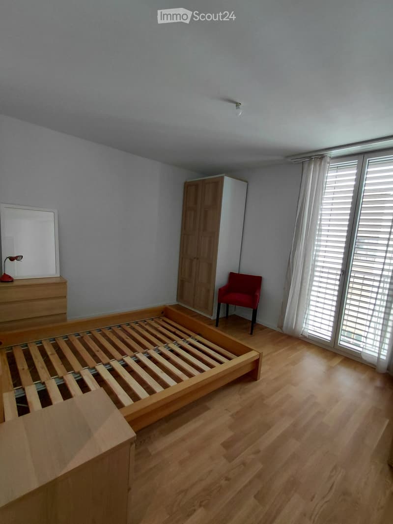Furnished Room. Bedroom with wooden floor, large window with blinds, bed frame, red chair, wardrobe, lamp, and mirror.