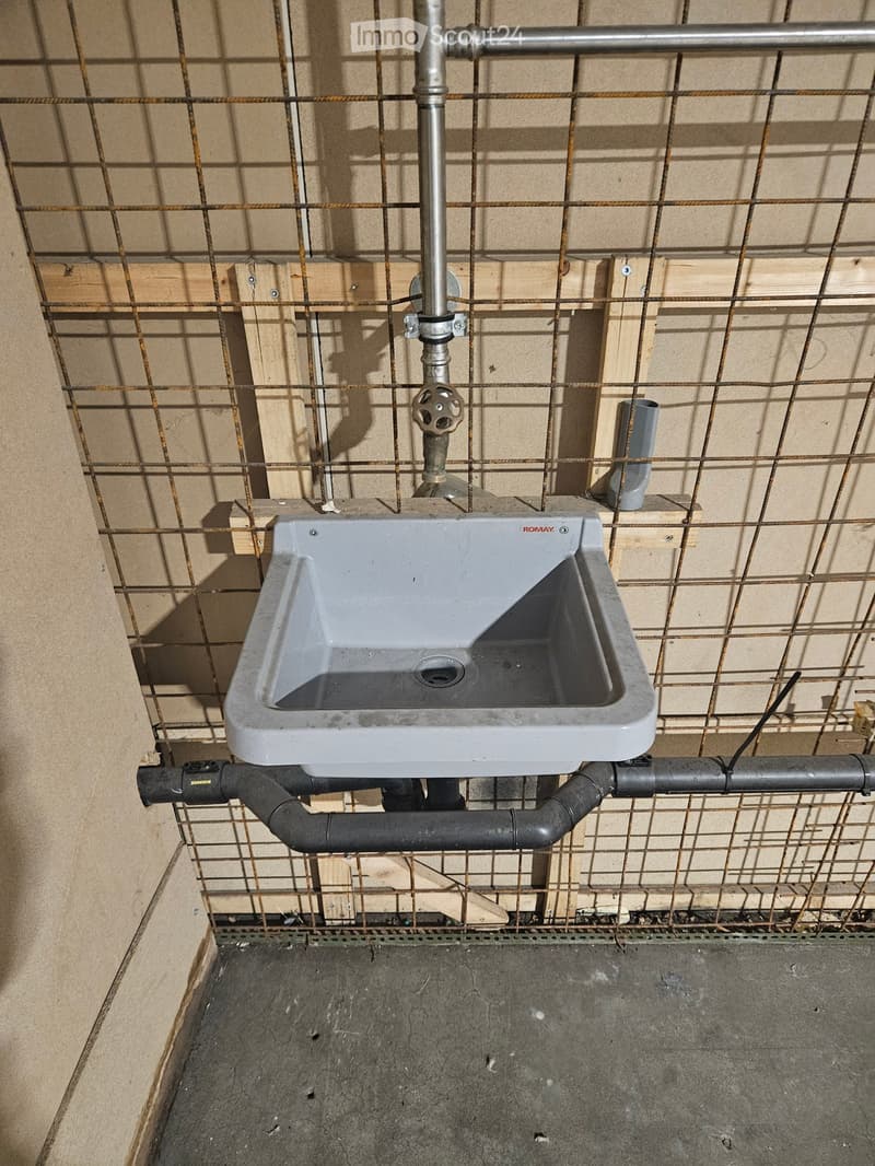 Bathroom. A urinal mounted on a concrete floor with visible plumbing pipes and a metal valve above it. The surrounding area includes exposed wooden framing and metal mesh.