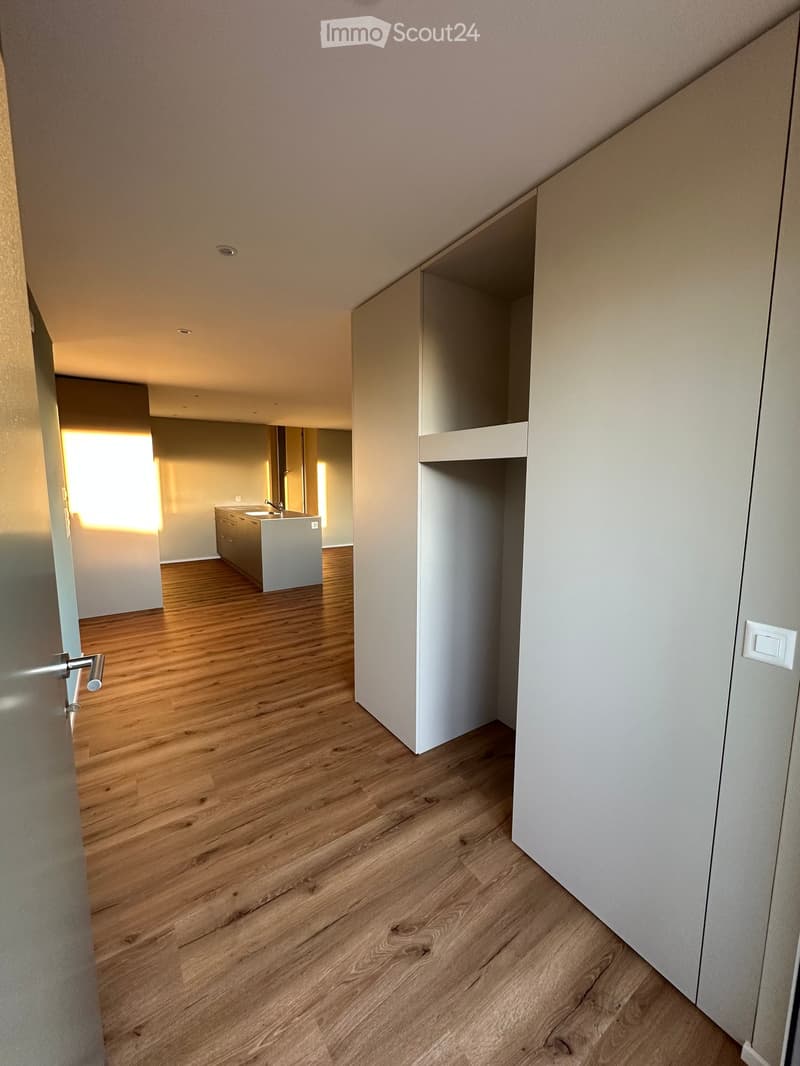 Corridor / Hallway. Empty hallway with wooden flooring, white walls, and a door leading to a kitchen area with cabinets and a sink.