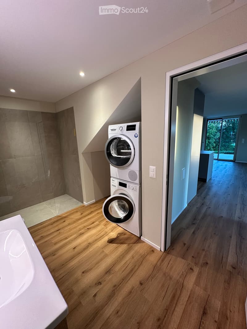 Furnished Room. Modern laundry room with wooden flooring, white walls, and built-in washing and drying machines.