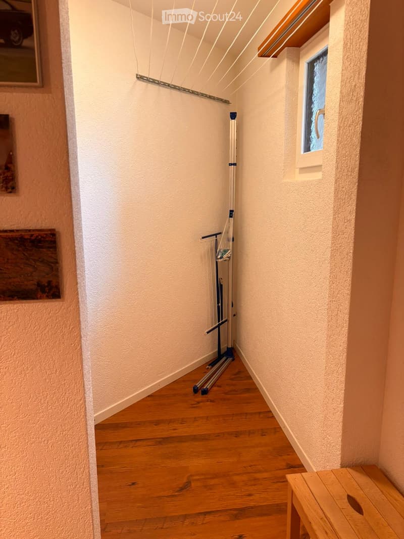 Corridor / Hallway. A narrow hallway with wooden flooring, white walls, and a small window on the right side. A clothes drying rack is positioned against the wall, and a small wooden bench is placed near the window.