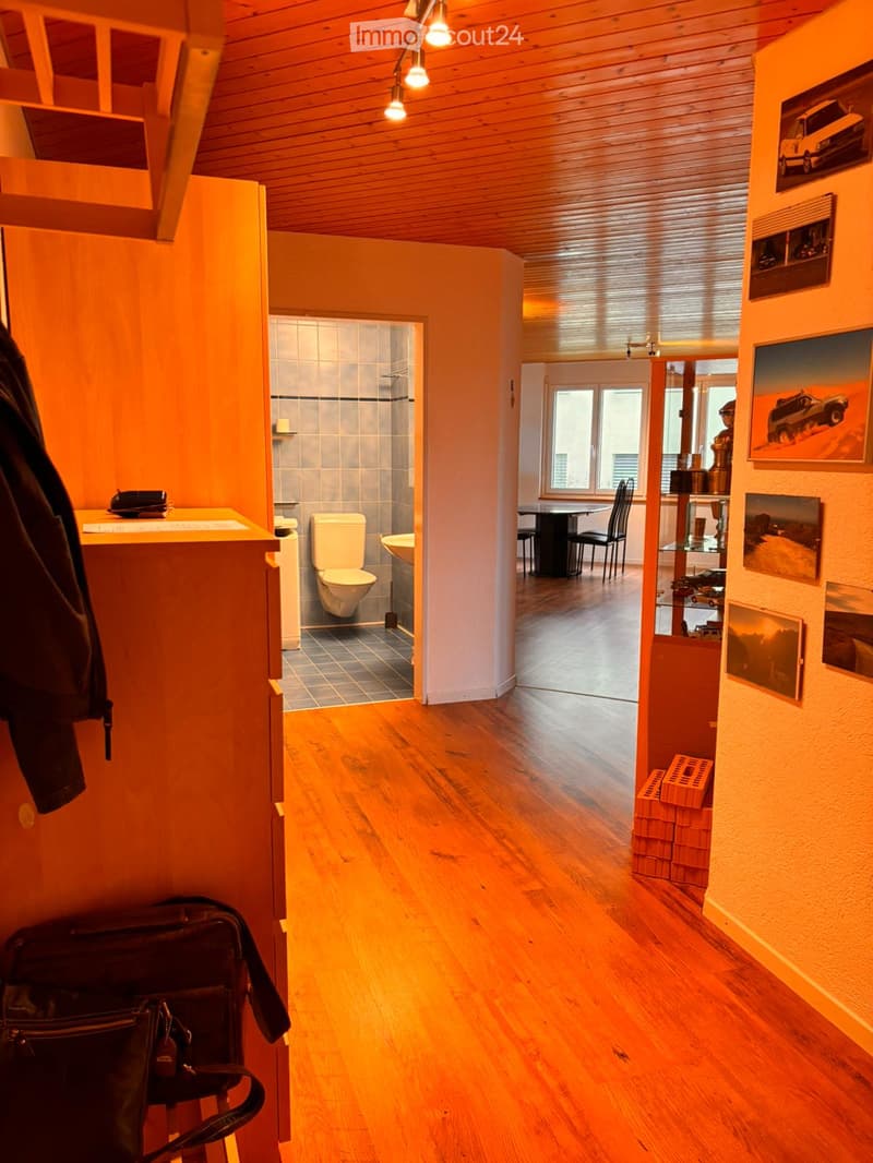 Corridor / Hallway. A well-lit hallway with wooden flooring, leading to a bathroom and a dining area. The hallway features artwork on the walls and a small stack of bricks on the floor.