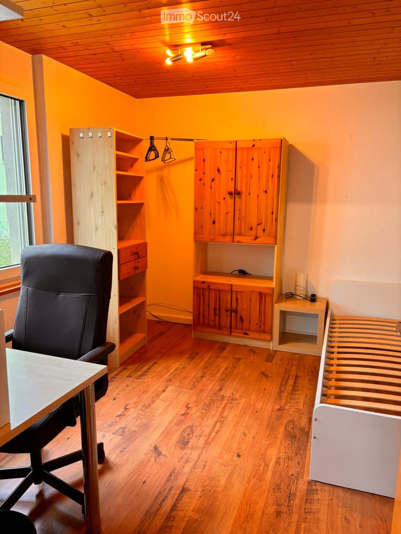 Furnished Room. A cozy, well-lit room with wooden flooring, a large window, a black office chair, a wooden table, and wooden cabinets with shelves.