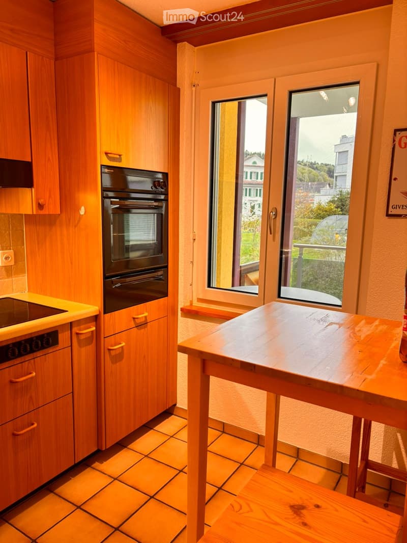 Kitchen. Bright kitchen with wooden cabinets, tiled floor, and a wooden table near large windows offering a view of the outside.