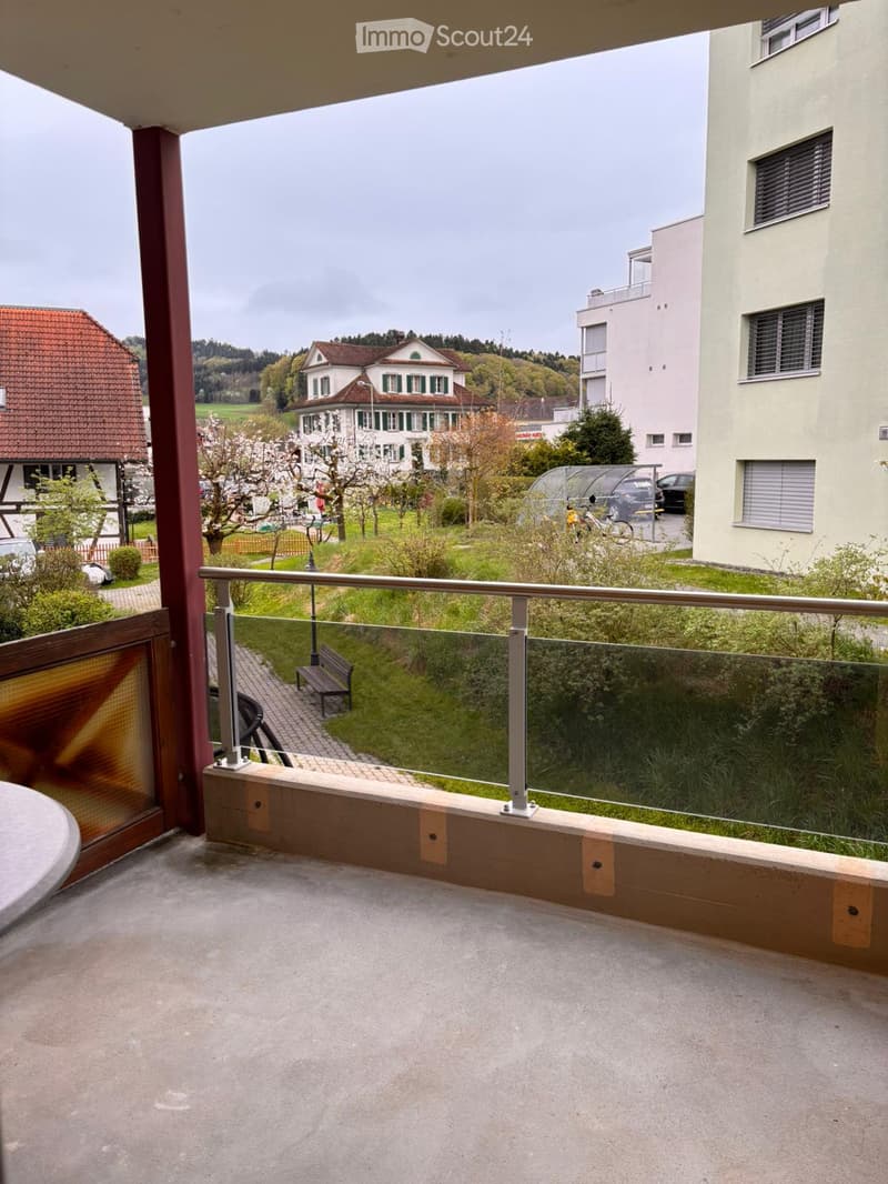 Balcony / Terrace. Balcony with glass railing, concrete floor, and view of a green park area with a bench, trees, and nearby buildings.