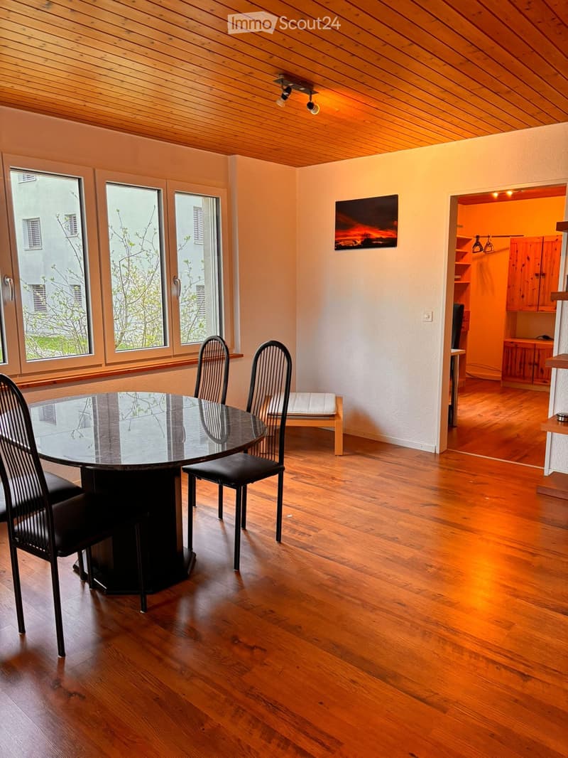 Furnished Room. Dining area with a round glass table, four black chairs, a wooden stool, and a small cabinet with shelves.