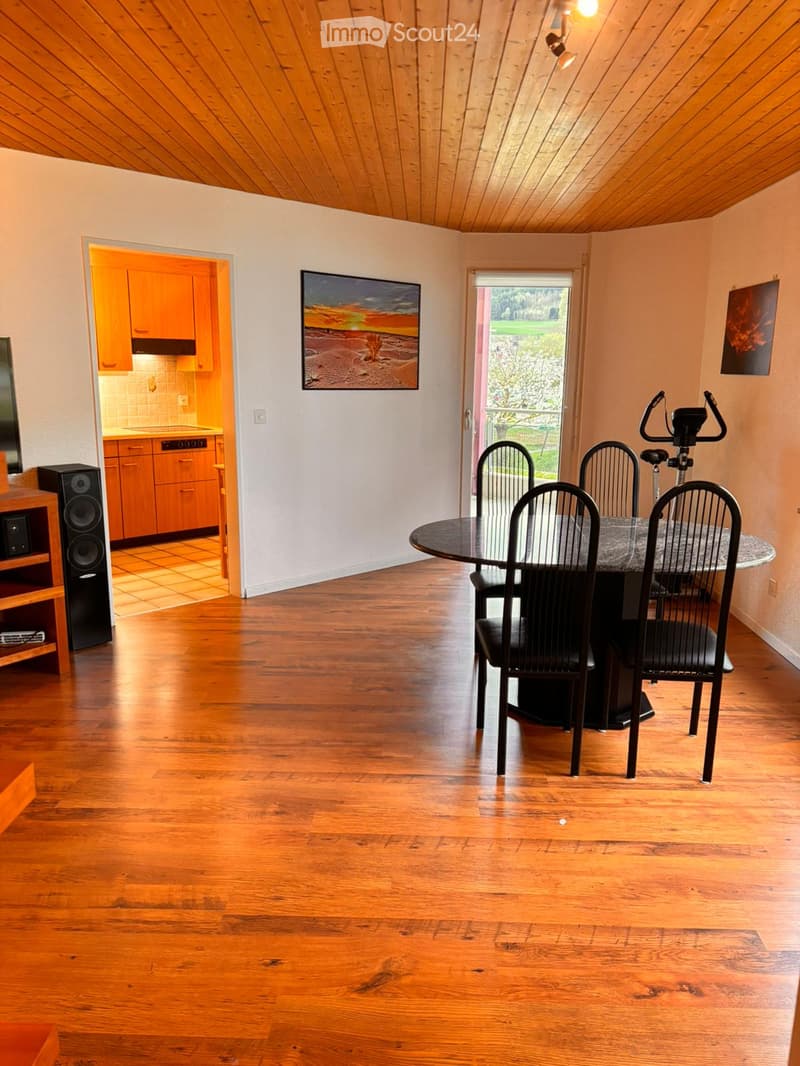 Furnished Room. Dining area with wooden flooring, a round dining table with four black chairs, a speaker system, an exercise bike, and a view of a kitchen area with wooden cabinets.