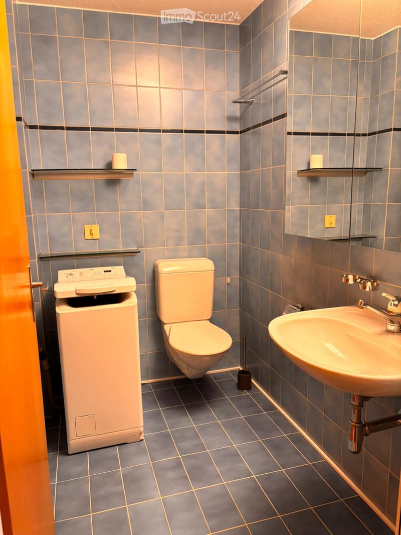 Bathroom. Modern bathroom with blue tiled walls, a toilet, a sink with a mirror, a towel rack, and a shelf holding a candle. The floor is tiled in dark grey with a subtle pattern.