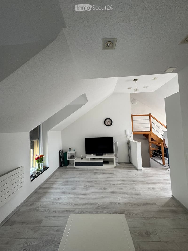 Living Room. Modern living room with light grey wooden flooring, white walls, and a large window. Features a TV on a white stand, a small side table with a vase of flowers, and a staircase leading to an upper level.