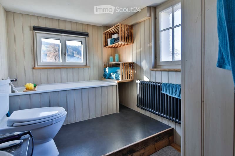 Bathroom with a toilet, bathtub, radiator, wooden shelves holding towels, and a view of a snowy landscape through the windows.