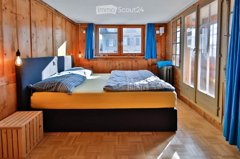 Cozy bedroom with wooden walls and flooring, featuring a large bed with blue and yellow bedding, a nightstand, and blue curtains on the windows.