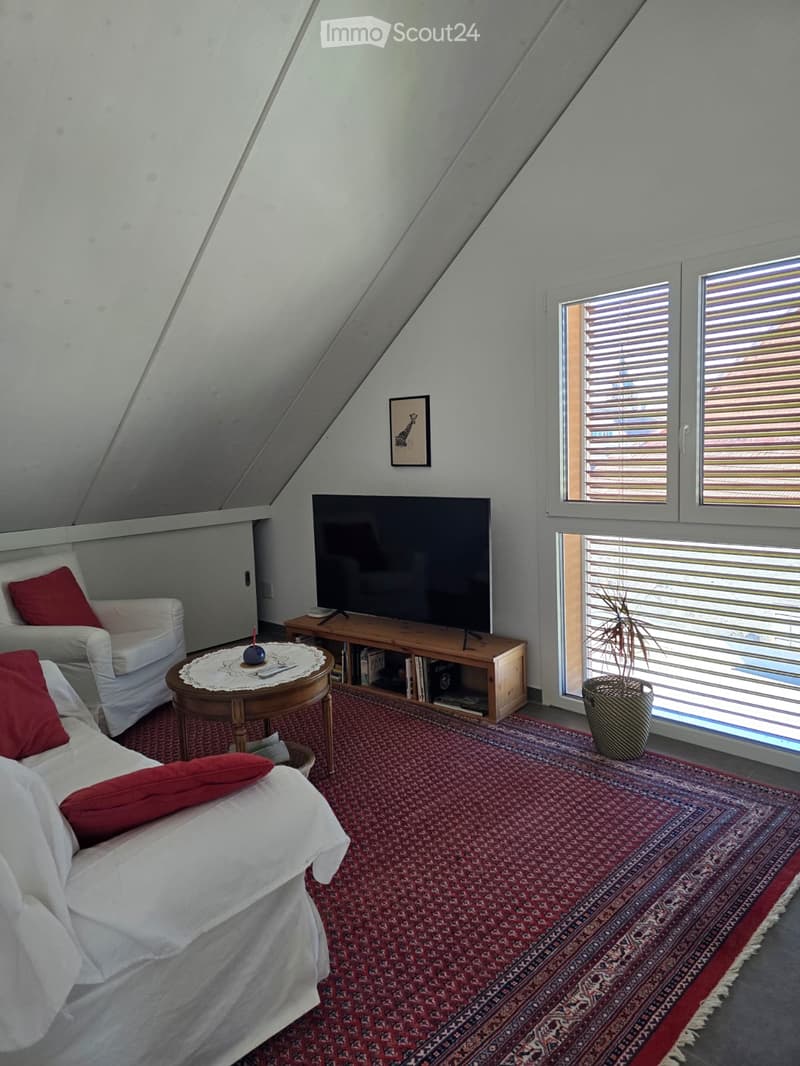Furnished Room. Bright attic living room with slanted ceilings, large windows, red patterned rug, white sofa, armchair, coffee table, TV, and decorative plant.