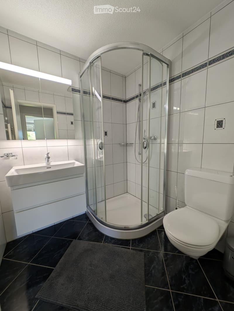 Bathroom. Modern bathroom with white tiles, a glass-enclosed shower, a white sink, and a toilet. Features a large mirror above the sink and a dark tiled floor.