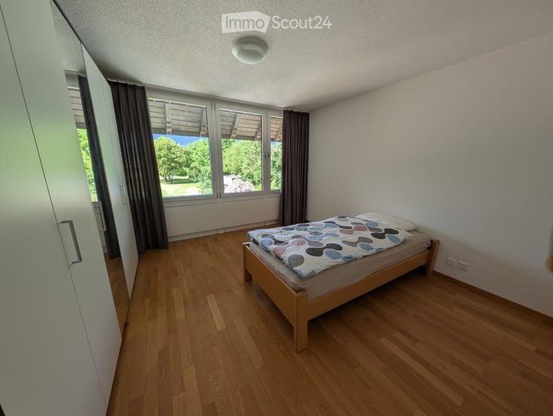Bedroom. Minimalist bedroom with wooden flooring, a single bed with a colorful quilt, and a large window with curtains offering a view of greenery.