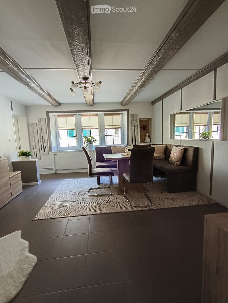 Furnished Room. Modern living room with exposed wooden beams, large windows, a sofa, dining table, chairs, and decorative elements.