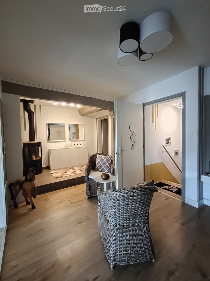 Furnished Room. A modern, well-lit living space with wooden flooring, a wicker chair, a small table with a pillow, a fireplace, and a cabinet with mirrors in the background.