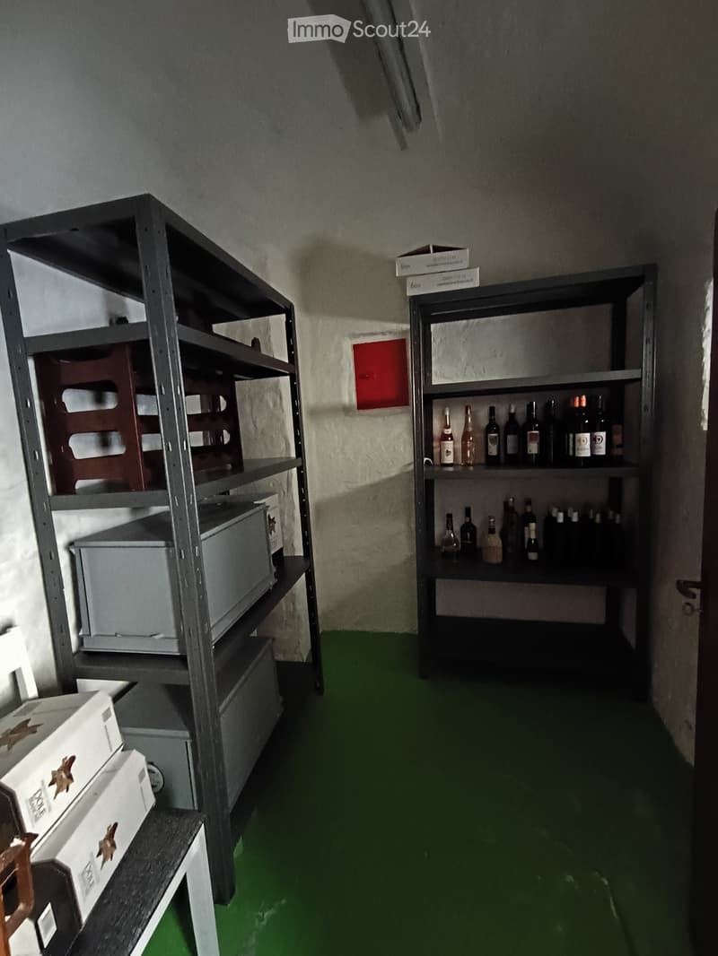 Furnished Room. A small, furnished room with green flooring and gray walls. It features two large metal shelves holding various items, including wine bottles and boxes, and a small table with additional boxes on it.