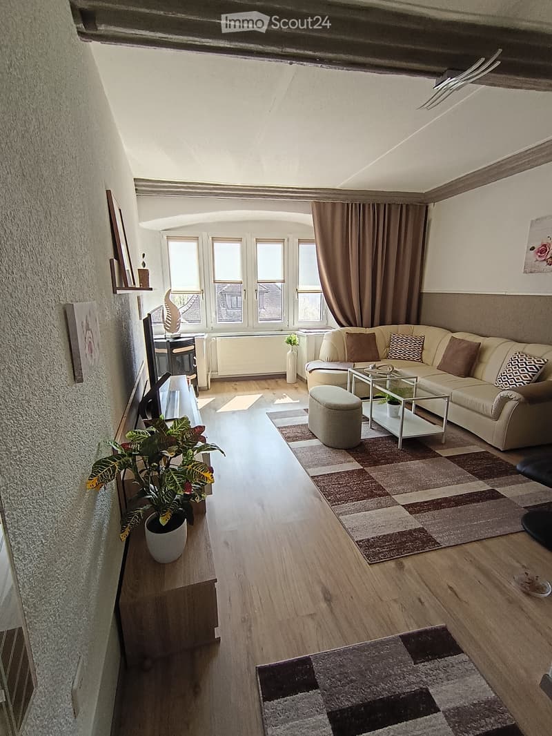 Living Room. Bright living room with wooden flooring, large windows, cream-colored sofa, glass coffee table, ottoman, and potted plants.