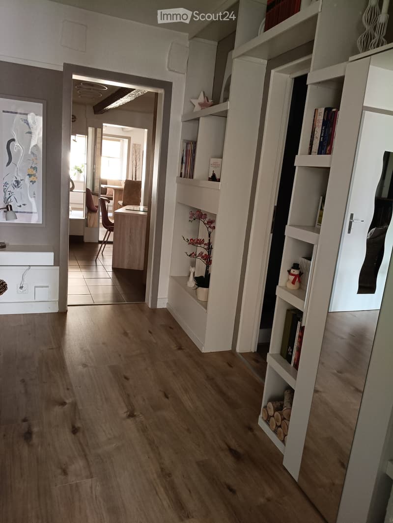 Corridor / Hallway. Hallway with wooden flooring, built-in shelving, and a small decorative plant on a shelf. The hallway leads to an adjacent room with a desk and chairs.