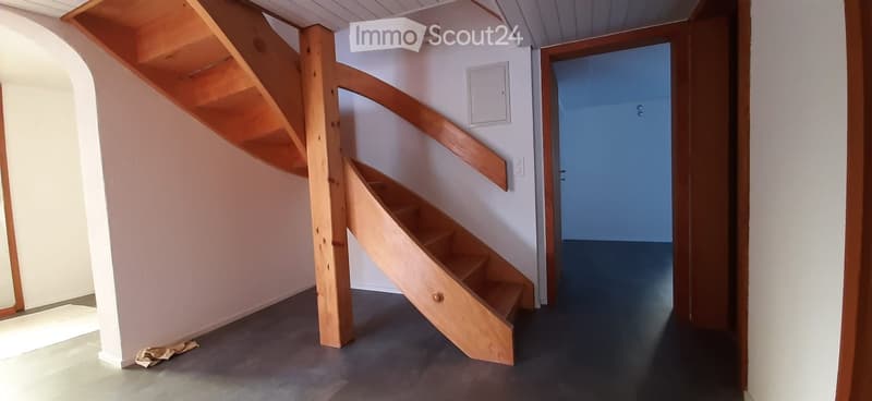 Corridor / Hallway. Spacious hallway with a wooden staircase leading upwards, white walls, and a blue door at the end.