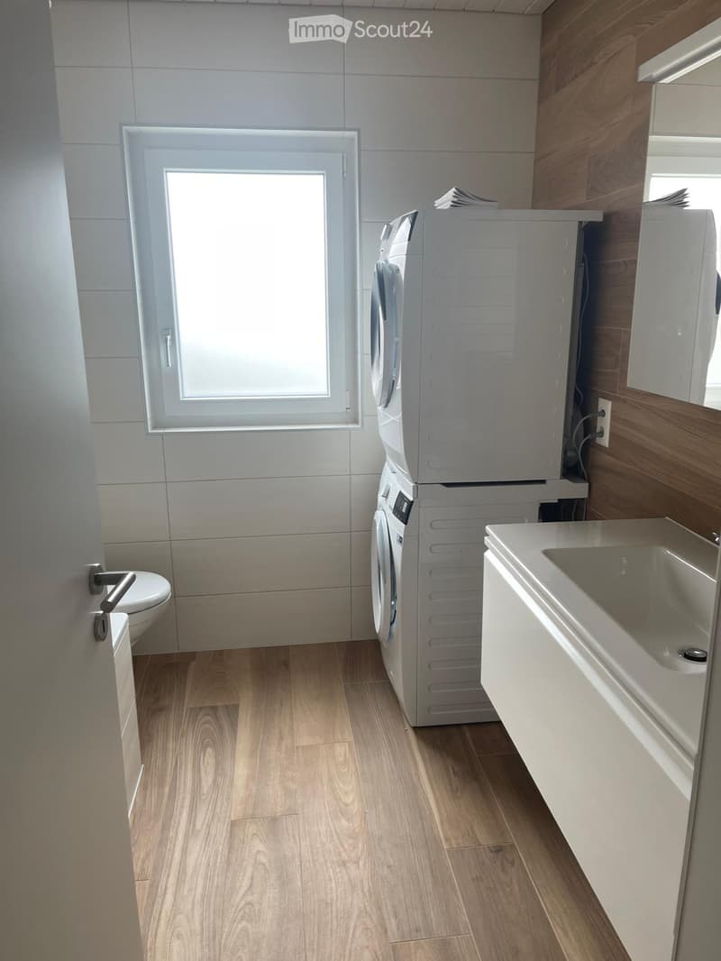 Bathroom. Bathroom with wooden flooring, white tiled walls, a toilet, a washing machine, a sink with a cabinet, and a window providing natural light.
