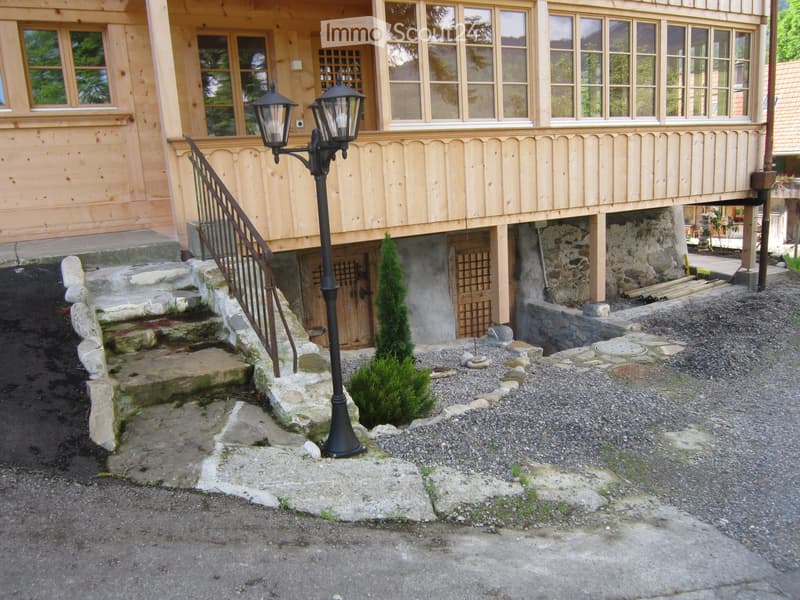 House. Two-story wooden house with large windows, stone steps leading to the entrance, and a small garden area with gravel and a small tree.