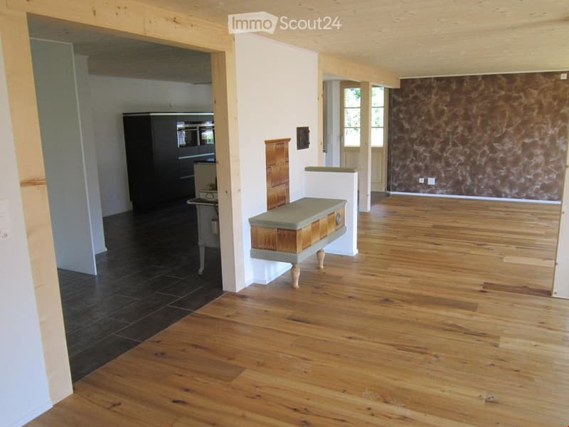 Unfurnished Room. Spacious, open-plan area with wooden flooring, white walls, and a mix of dark and light flooring. Features include a small table, a bench, and a kitchen area visible in the background.