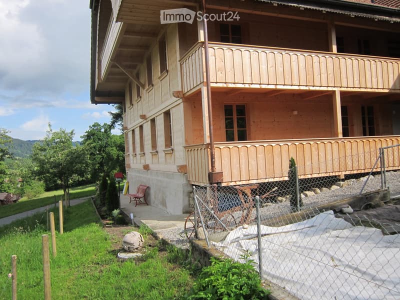 House. Two-story wooden house with a balcony, surrounded by greenery and a fenced area.