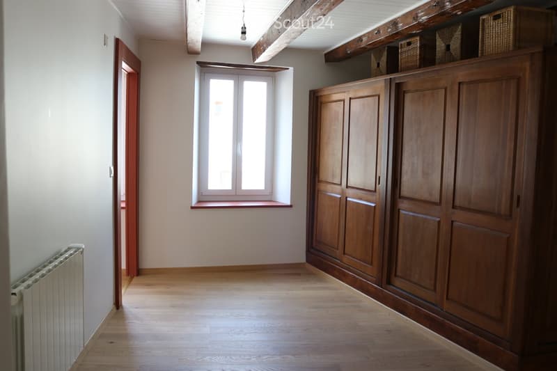 Unfurnished Room. Empty room with wooden flooring, white walls, large window with wooden sill, built-in wooden cabinetry, exposed wooden beams on the ceiling, and a radiator on the left side.