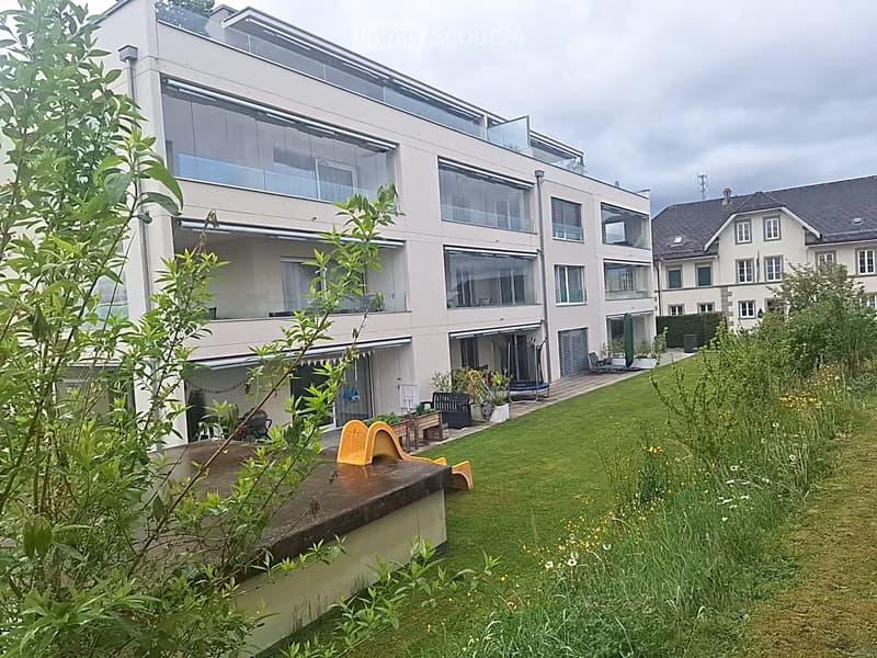 Building. Modern multi-story building with large windows and a spacious garden area featuring a wooden deck, lounge chairs, and a small play structure.