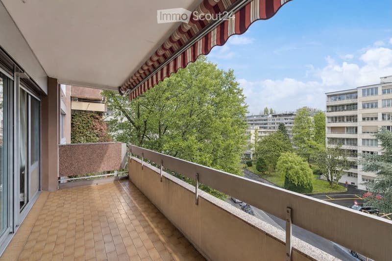 Balcony / Terrace. Balcony with tiled floor, metal railing, striped awning, overlooking a green area with trees and buildings.
