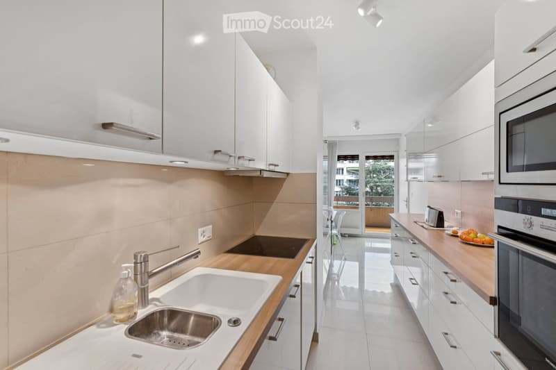 Kitchen. Modern kitchen with white cabinets, wooden countertops, stainless steel sink, and appliances including an oven, microwave, and toaster. A fruit bowl is on the counter, and a view of a balcony is visible through the glass door.
