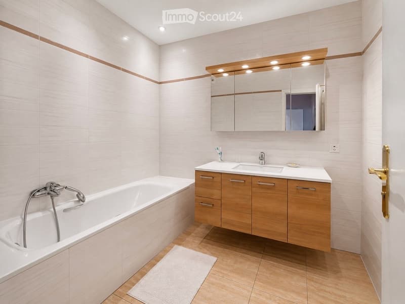 Bathroom. Modern bathroom with a white bathtub, wooden bathroom cabinet, sink, and large mirror with integrated lighting. The walls are tiled in light beige with brown accents, and the floor is wooden.