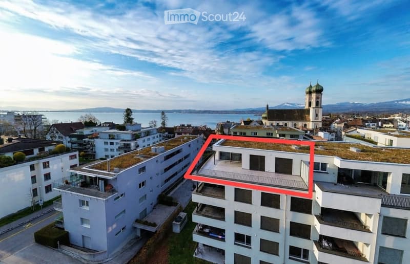 Building. Aerial view of a modern residential complex with multiple buildings, green roofs, balconies, and a scenic backdrop of a lake and distant mountains.