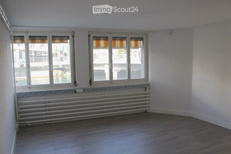 Empty room with wooden flooring, white walls, large windows with blinds, and a radiator below the windows.